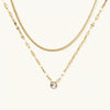 Layered gold necklace with herringbone and link chains, small bezel-set crystal pendant on cream background